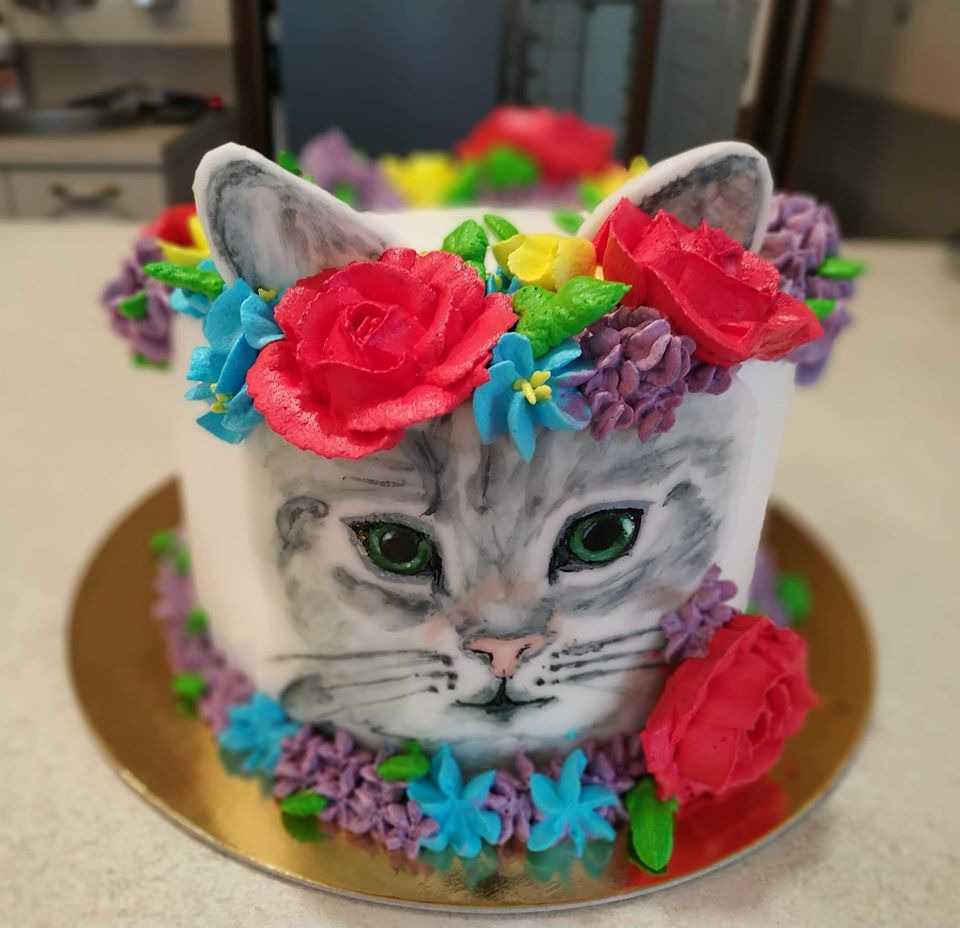 Tort z ręcznie malowaną twarzą szarego kota z zielonymi oczami, otoczony kremowymi kwiatami w odcieniach czerwieni, fioletu i błękitu, umieszczony na złotej podkładce.