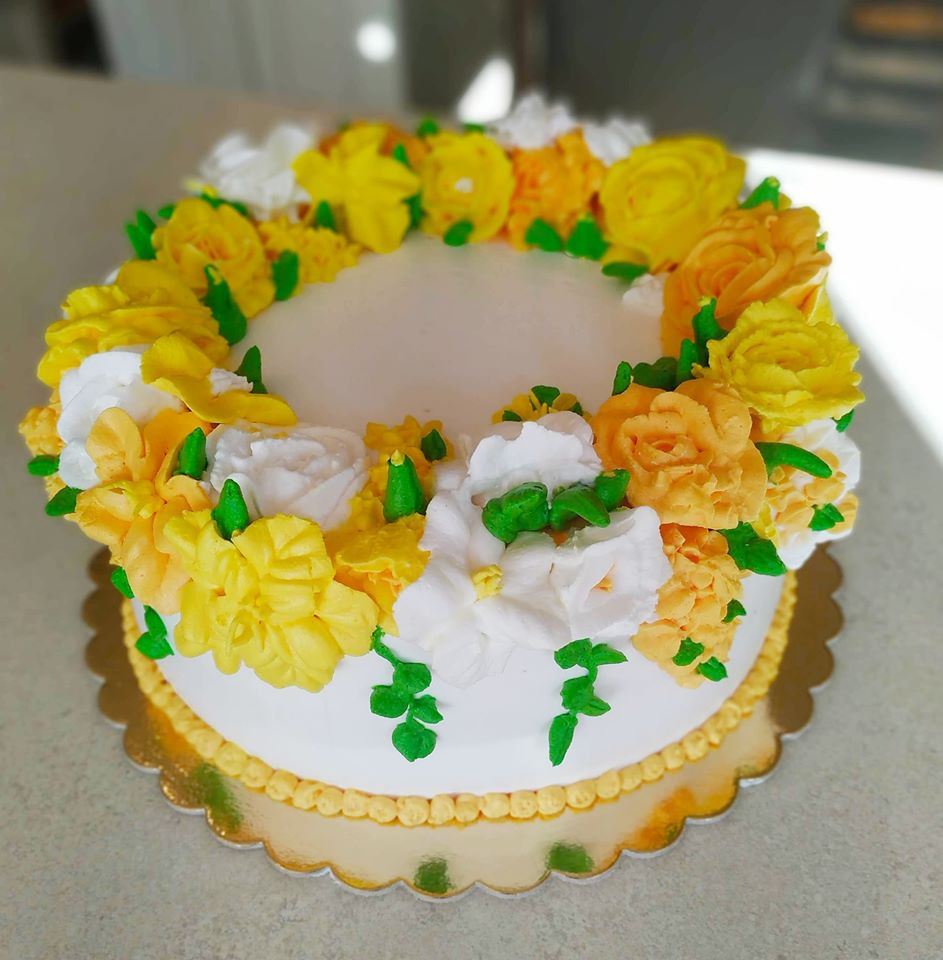 Tort z białym kremem i dekoracją z kremowych kwiatów w odcieniach żółci i bieli, umieszczony na złotej podkładce.