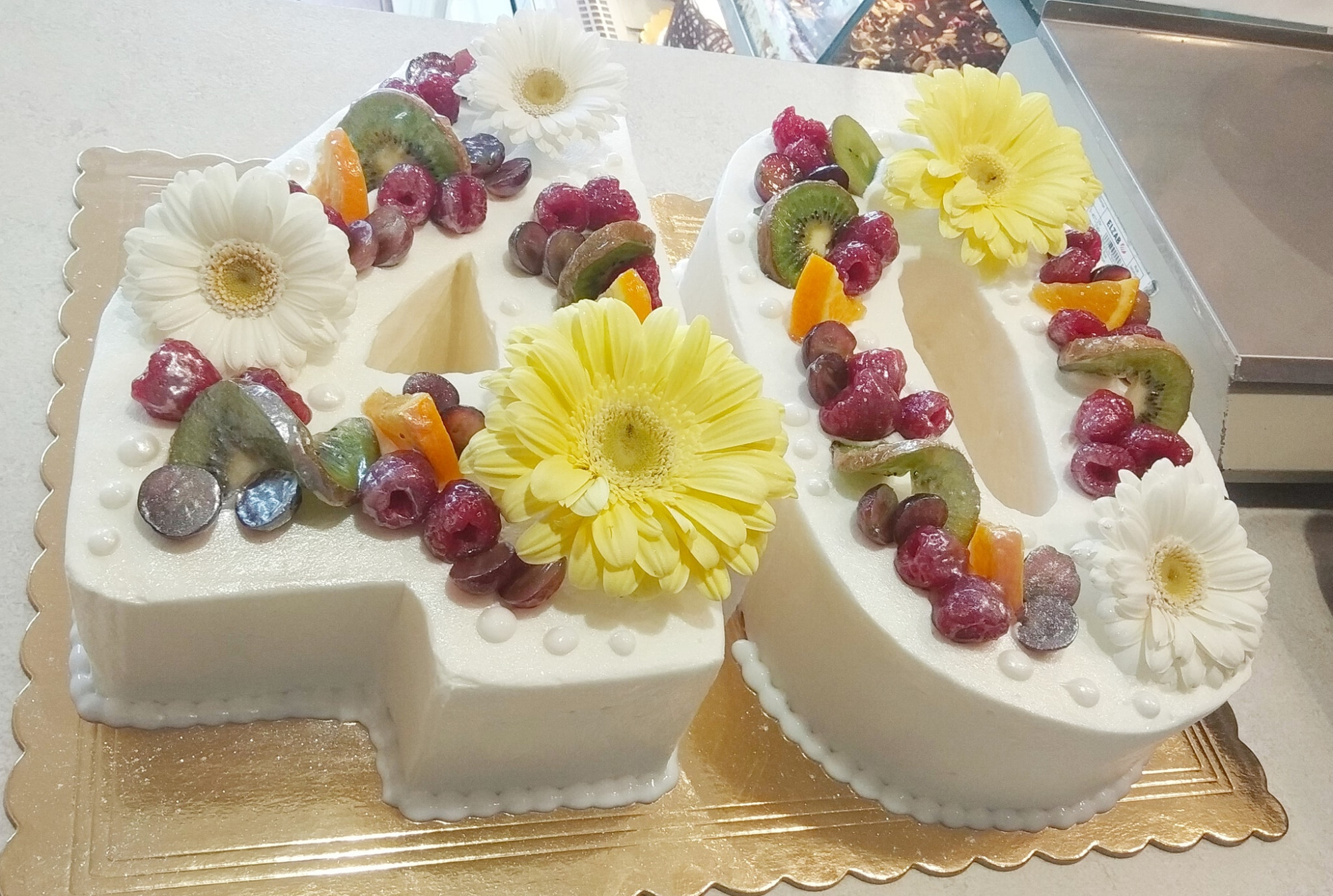 Tort w kształcie liczby 40, udekorowany białym lukrem, świeżymi malinami, kiwi, mandarynkami, winogronami i białymi oraz żółtymi gerberami, na złotej podkładce.