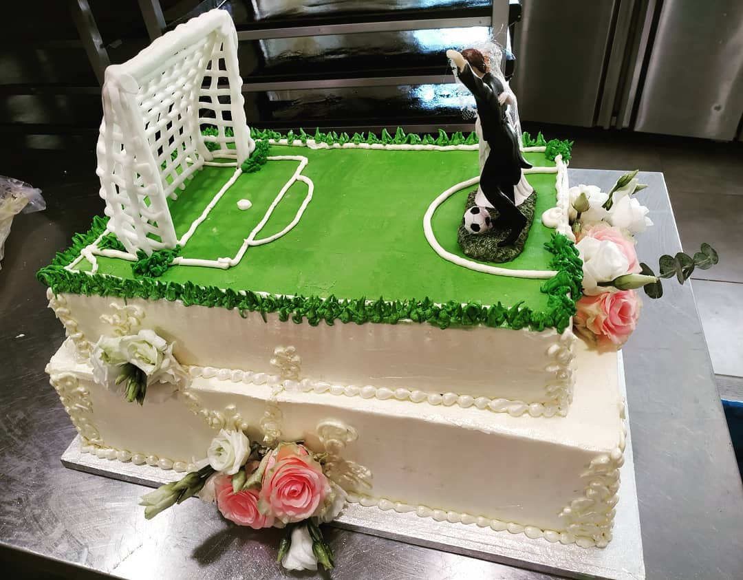 Dwupoziomowy tort weselny z motywem piłkarskim: zielone boisko z bramką z białej czekolady, figurka pary młodej z piłką i dekoracje z kremowych róż.