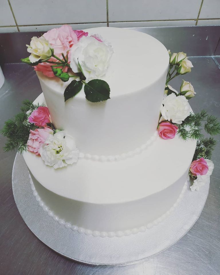 Dwupiętrowy tort weselny z białym lukrem, ozdobiony różowymi i białymi kwiatami oraz drobnymi zielonymi gałązkami, umieszczony na srebrnej tacy.