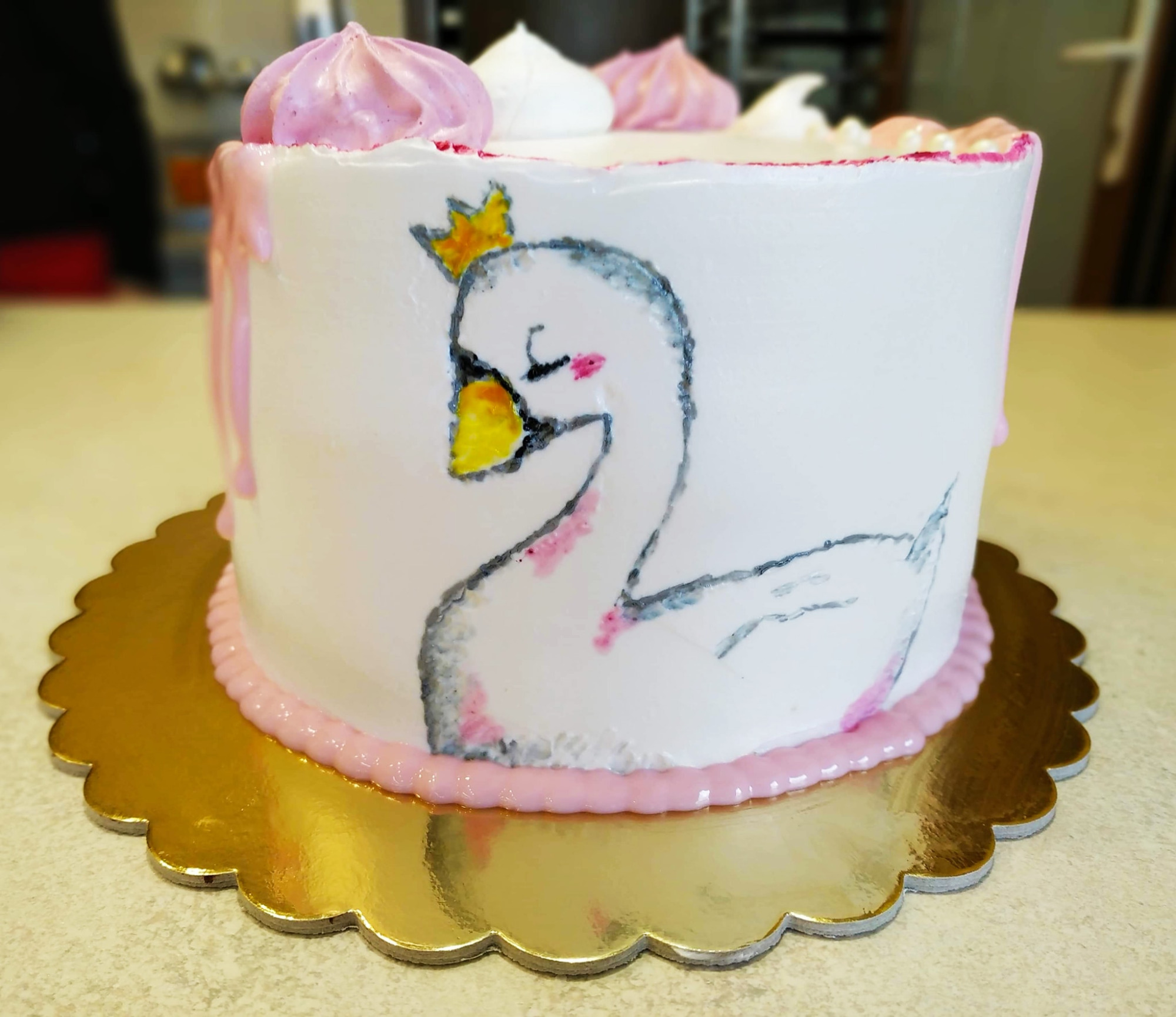 Tort z białym lukrem, dekorowany rysunkiem śpiącego łabędzia w koronie i różowymi bezami na wierzchu, umieszczony na złotej, ozdobnej podkładce.