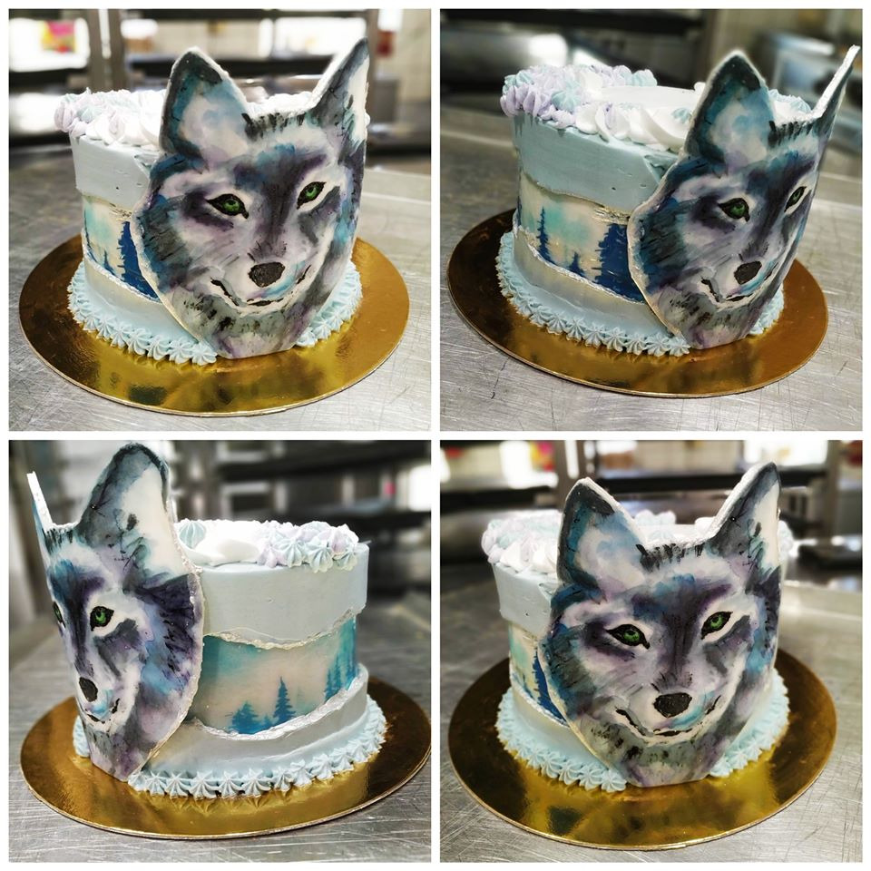 Tort zdobiony motywem wilka w odcieniach błękitu i szarości, z dekoracją imitującą zimowy krajobraz, umieszczony na złotej podkładce.