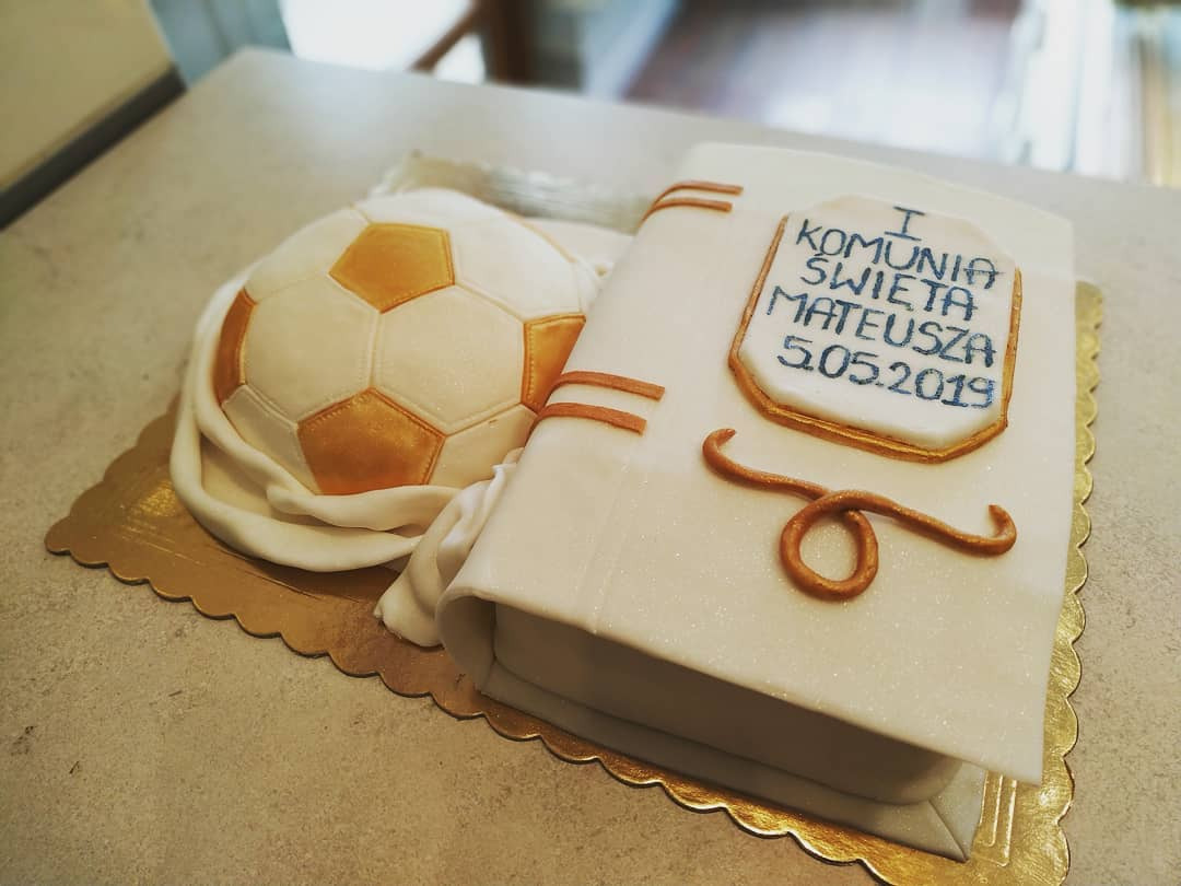 Tort komunijny w kształcie otwartej księgi i piłki nożnej, z napisem 'I Komunia Święta Mateusza 5.05.2019' w niebieskiej ramce i złotymi zdobieniami, na złotej podkładce.