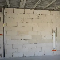 Ściana z betonu komórkowego z wyżłobieniami pod przewody elektryczne i rurę, widoczne pomarańczowe puszki instalacyjne, częściowo wypełnione pianką montażową.