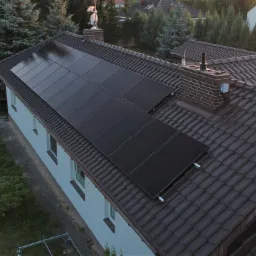 Ciemne panele słoneczne na brązowym, spadzistym dachu domu jednorodzinnego, widoczne okna i zielone drzewa w tle, ujęcie z góry.