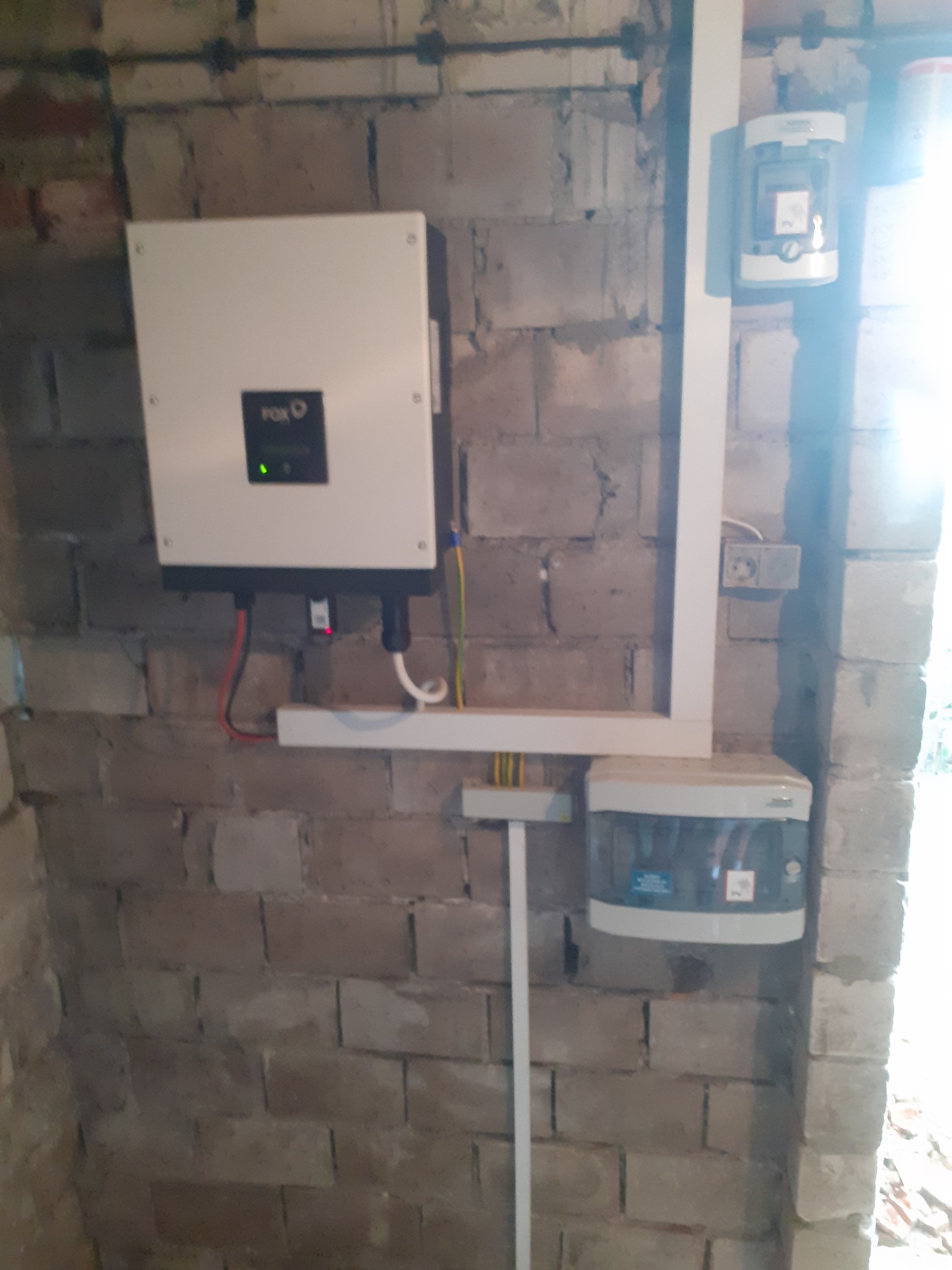 Zamontowany inwerter FoxESS na surowej ścianie z cegły, z widoczną instalacją elektryczną w białych listwach, skrzynka zabezpieczeń i licznik energii.