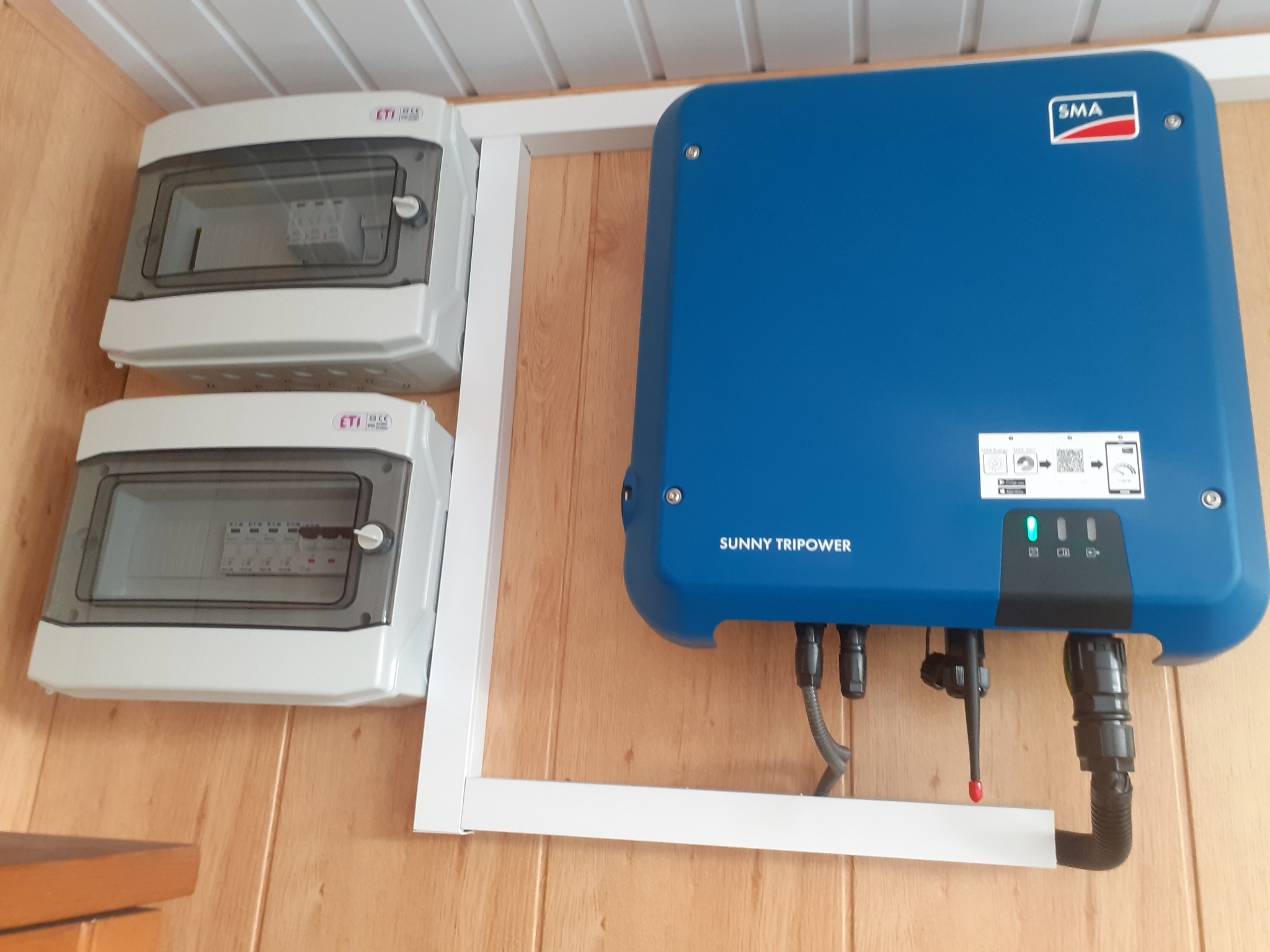 Niebieski inwerter fotowoltaiczny SMA Sunny Tripower zamontowany na drewnianej ścianie obok dwóch białych skrzynek bezpiecznikowych ETI