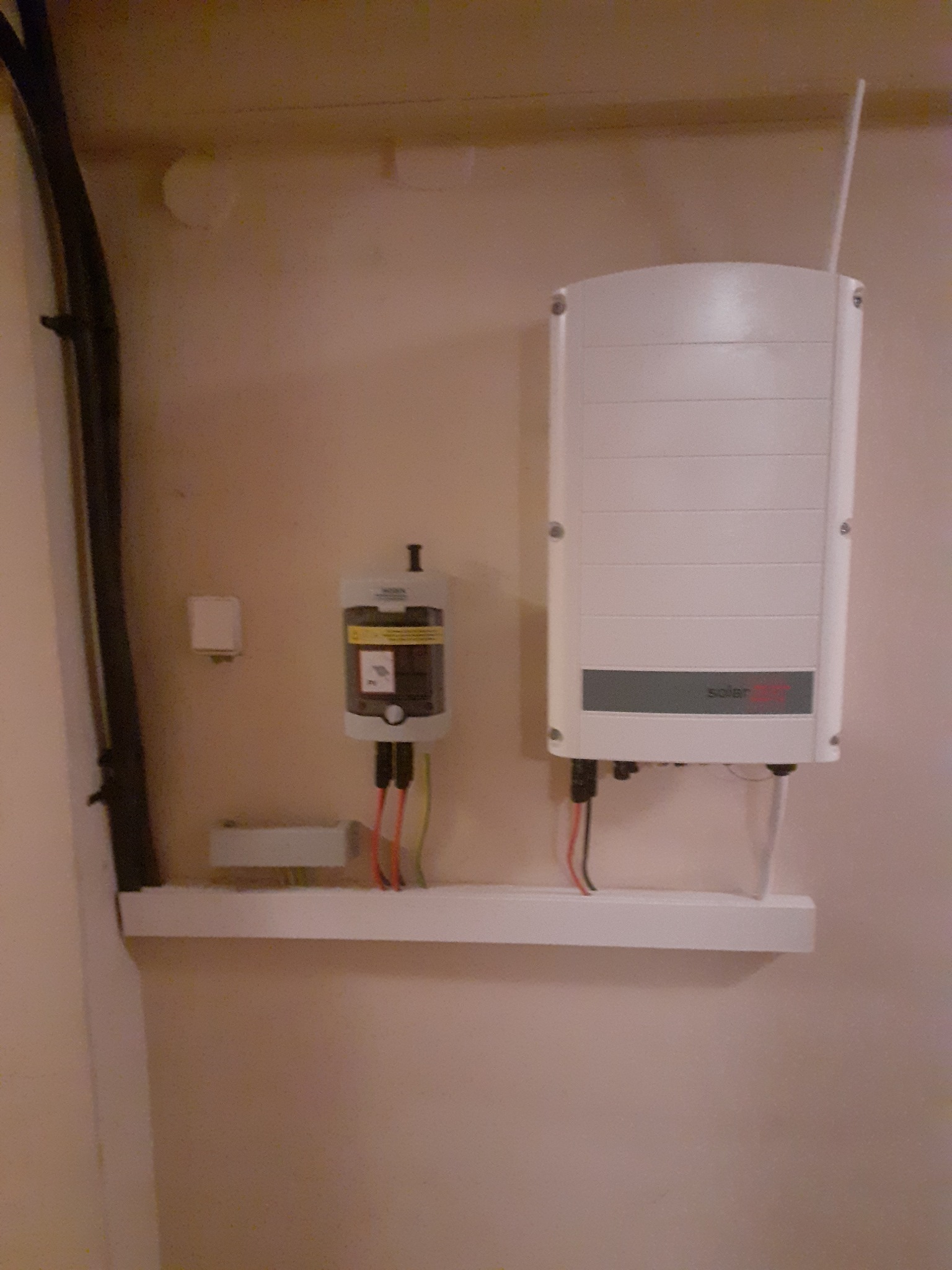 Zamontowany inwerter fotowoltaiczny SolarEdge z anteną i dodatkowe urządzenie na białej półce, widoczne kable zasilające.