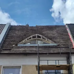 Częściowo zdemontowane pokrycie dachu z okrągłym oknem na poddaszu, widoczne rusztowanie i niebo z chmurami.