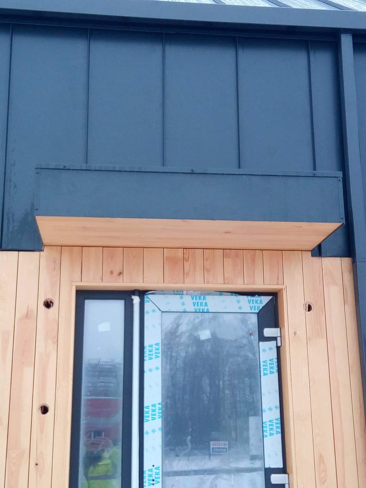 Fragment elewacji budynku z ciemną blachą na dachu i drewnianą okładziną ścian. Zamontowane okno z folią ochronną producenta VEKA. Widoczny fragment drewnianego okapu nad oknem.