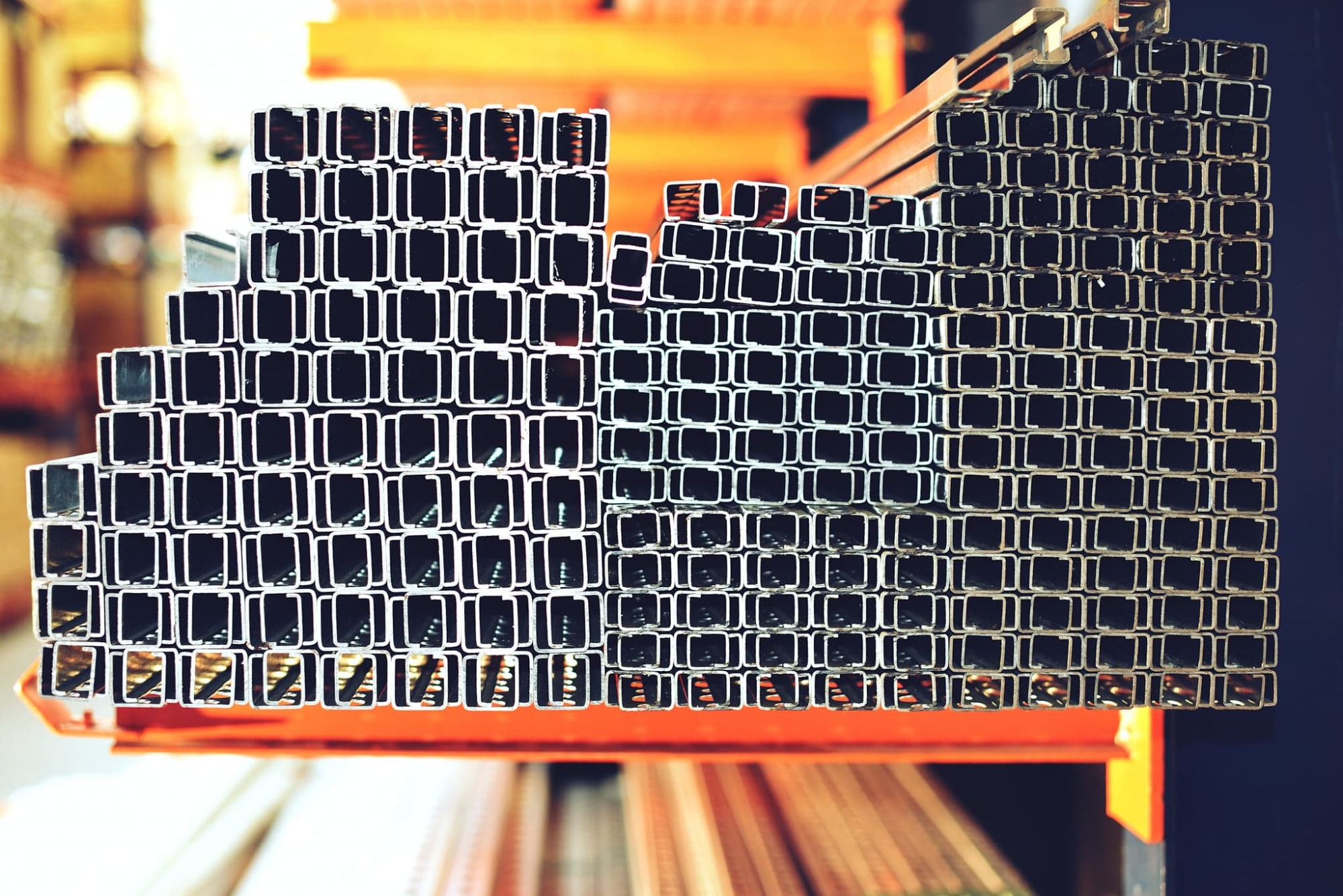 Stos metalowych profili kwadratowych ułożonych warstwami na pomarańczowym regale w magazynie.