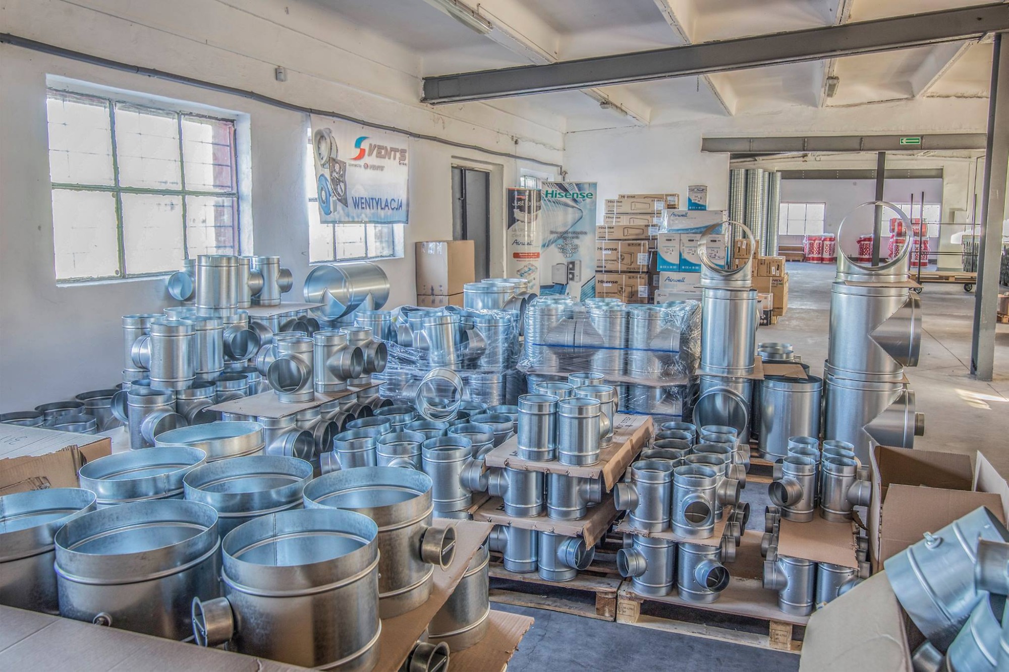 Wnętrze magazynu ze stertą ocynkowanych elementów wentylacyjnych, w tym rur, kolanek i trójników, ułożonych na paletach i w kartonach, w tle widoczny baner reklamowy i okno.