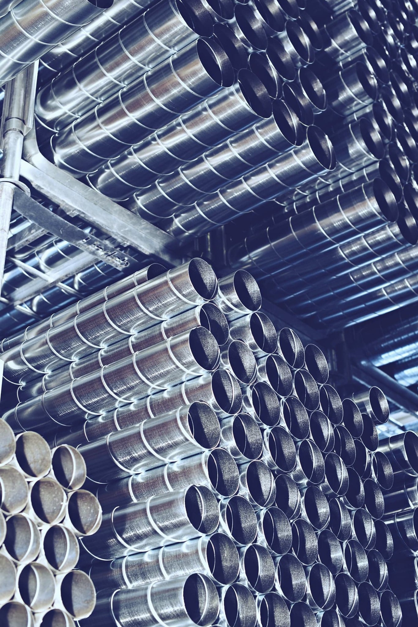 Stos spiralnych, metalowych rur wentylacyjnych ułożonych na półkach w magazynie, widok z dołu, oświetlenie sufitowe.