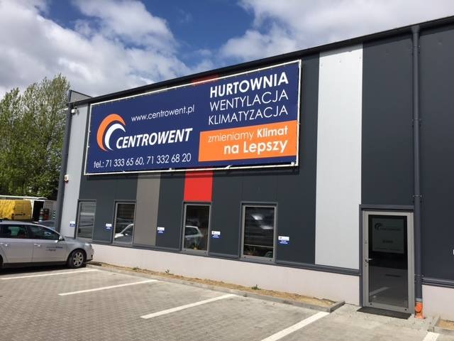 Budynek firmy Centrowent z dużym banerem reklamowym na fasadzie, informującym o hurtowej sprzedaży systemów wentylacji i klimatyzacji; widoczny parking i zaparkowany samochód.