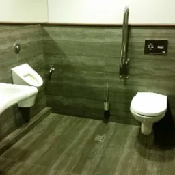 Toaleta publiczna z umywalką, pisuarami i uchwytami dla niepełnosprawnych, wykończona szarą płytką imitującą kamień, oświetlona światłem dziennym.