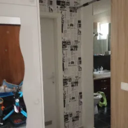 Wnętrze przedpokoju z tapetą imitującą gazety, widok na łazienkę z toaletą i umywalką, na pierwszym planie fragment szafy i wózek dziecięcy.