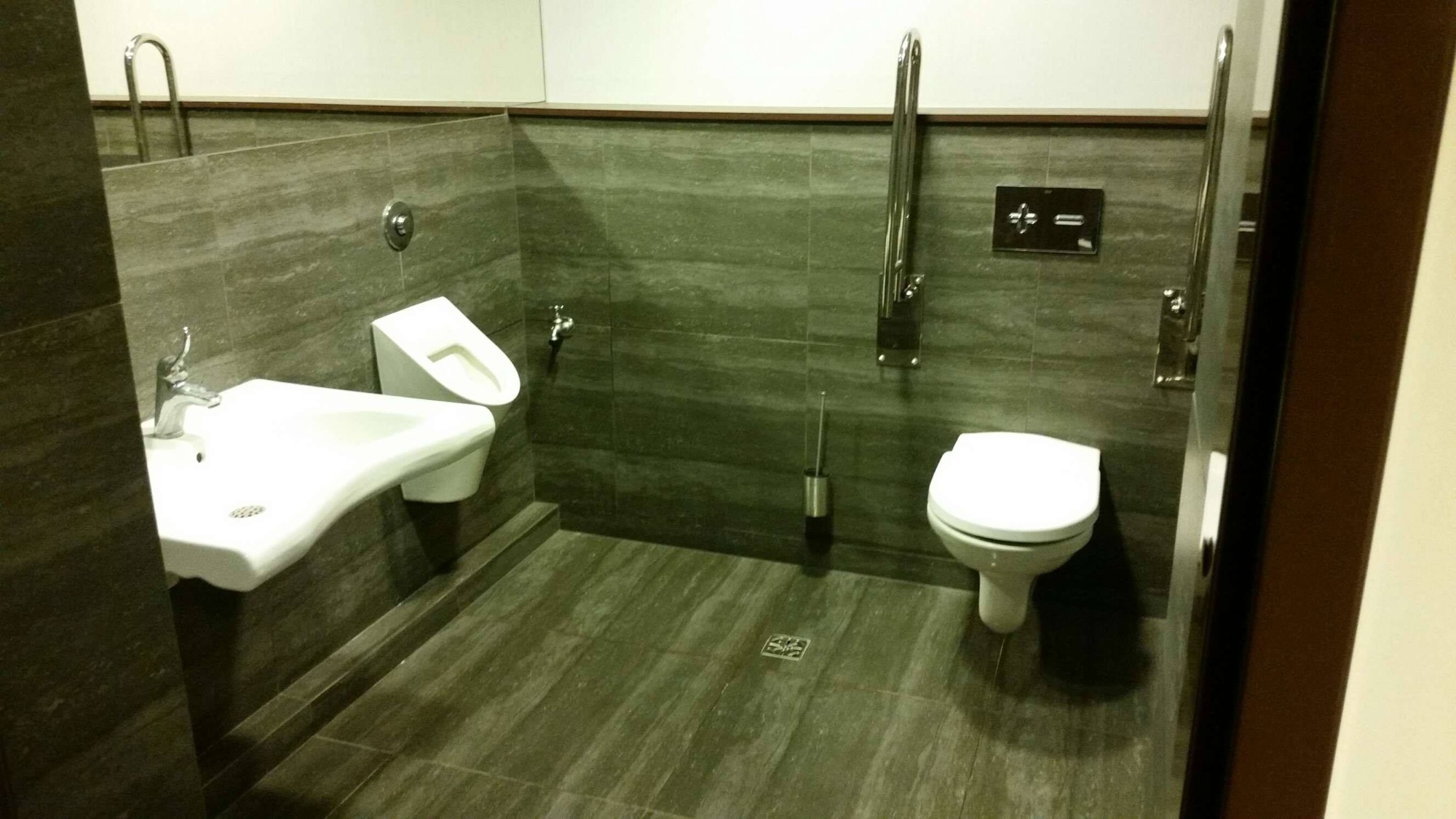 Toaleta publiczna z umywalką, pisuarami i uchwytami dla niepełnosprawnych, wykończona szarą płytką imitującą kamień, oświetlona światłem dziennym.