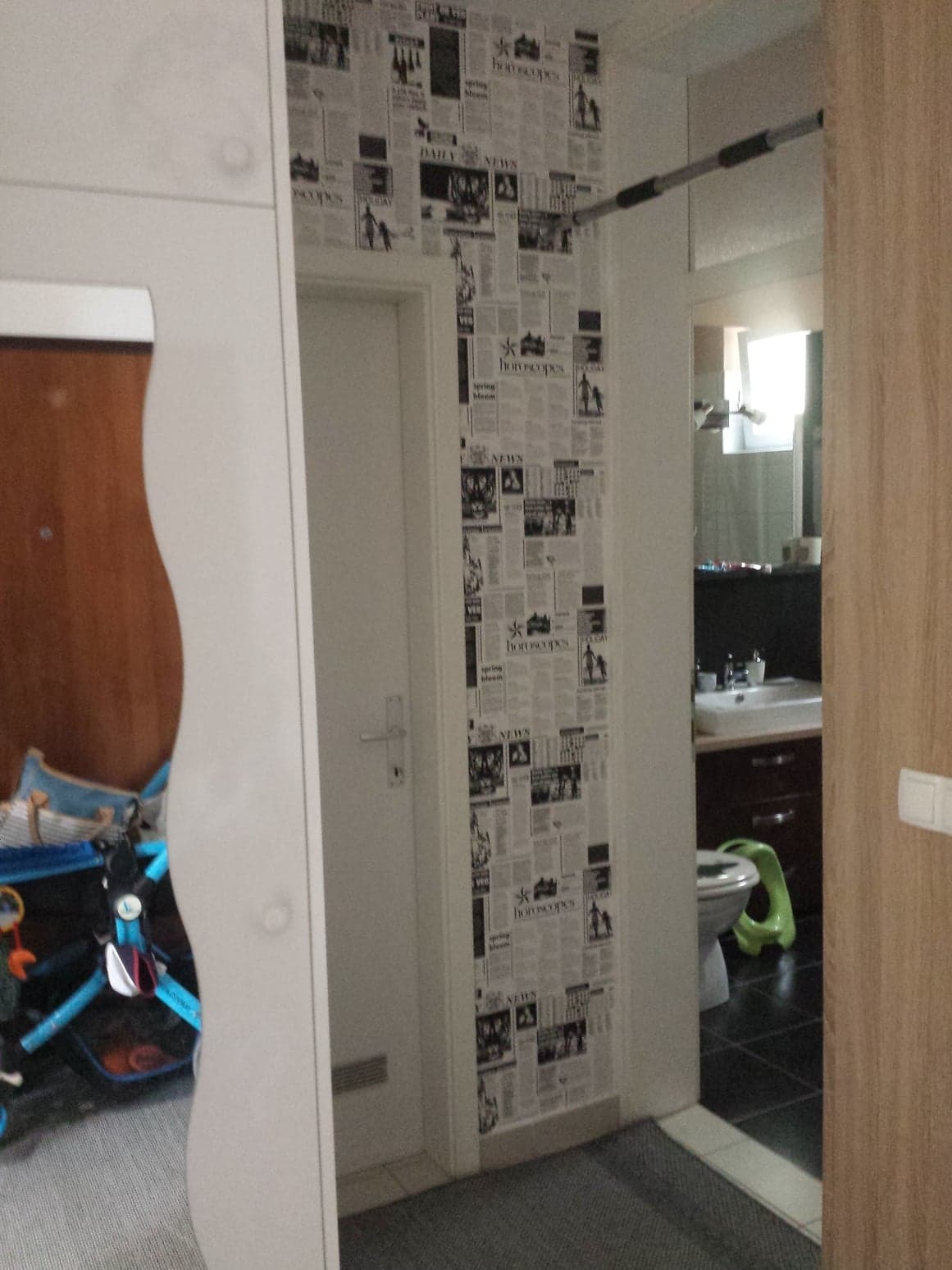 Wnętrze przedpokoju z tapetą imitującą gazety, widok na łazienkę z toaletą i umywalką, na pierwszym planie fragment szafy i wózek dziecięcy.