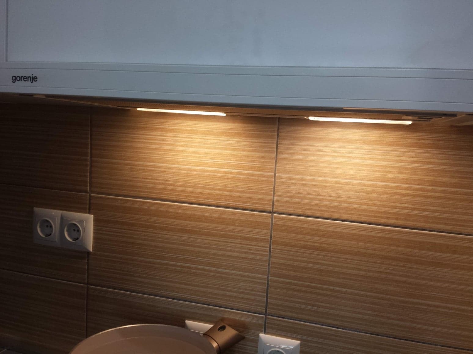 Kuchnia po remoncie: brązowe płytki imitujące drewno na ścianie nad blatem, oświetlenie podszafkowe LED, widoczne gniazdka elektryczne i fragment okapu Gorenje.