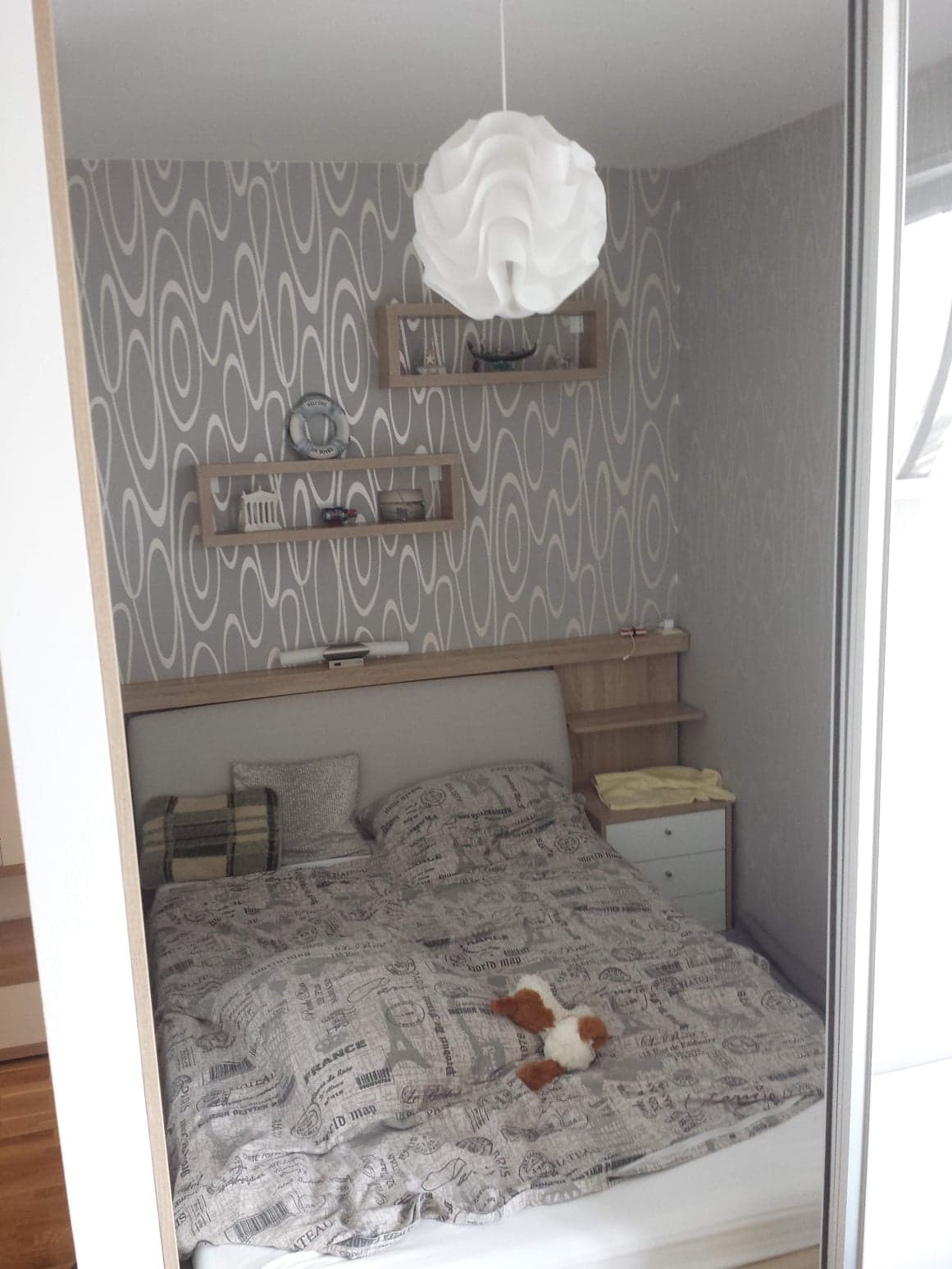 Sypialnia po remoncie: łóżko z szarą pościelą w stylu vintage, wzorzysta tapeta w odcieniach szarości, biała lampa wisząca, drewniane półki z dekoracjami.