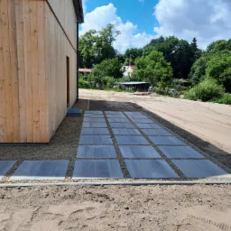 Nowoczesna ścieżka z szarych płyt betonowych ułożona na żwirze przed drewnianym domem w Wałczu, z widocznymi śladami opon na piasku.