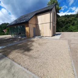 Nowoczesny dom z drewnianą elewacją i szarym dachem, widok z poziomu gruntu na podwórze z wysypanym żwirem i pojedynczymi płytami chodnikowymi, drabina oparta o ścianę budynku.