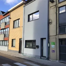 Wąska, pomalowana na szaro kamienica z dwoma oknami i czarną skrzynką na listy, widoczna z poziomu ulicy z przejściem dla pieszych.