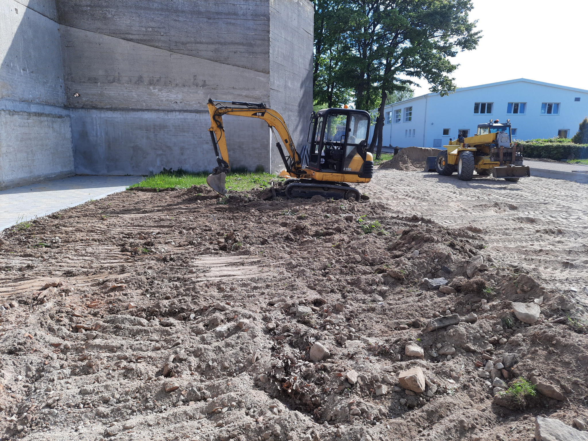 Żółta koparka CAT 303.5E w trakcie prac ziemnych na placu budowy, w tle budynek przemysłowy i ładowarka kołowa.