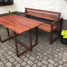 Drewniany stół i ławka z metalowymi nogami w kolorze brązowym, ustawione na brukowanym podwórku przed budynkiem z oknem z kwiatami.