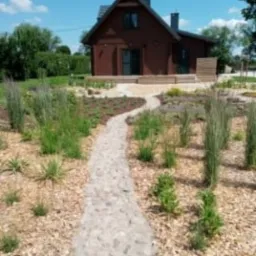 Kręta ścieżka z nieregularnych kamieni prowadząca przez rabatę z trawami ozdobnymi i korą drzewną w kierunku nowoczesnego domu z ciemnobrązową elewacją w słoneczny dzień.
