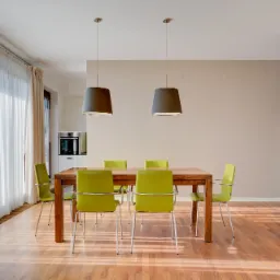 Jasne, przestronne wnętrze jadalni z drewnianym stołem, zielonymi krzesłami i dwoma lampami wiszącymi, widok na kuchnię w tle, parkiet z naturalnego drewna.