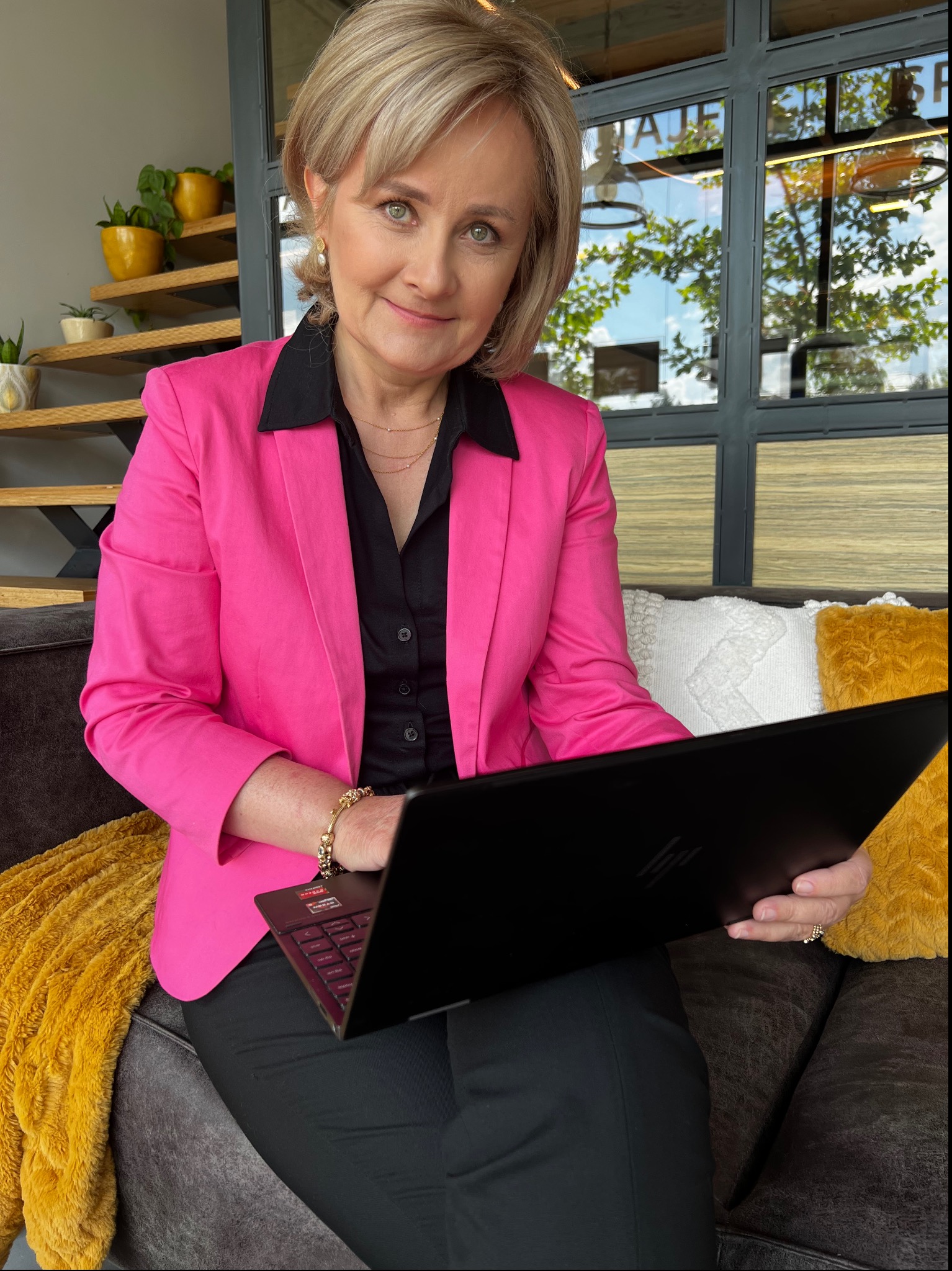 Kobieta w różowej marynarce i czarnej koszuli, siedząca na szarej kanapie z żółtym kocem i pracująca na czarnym laptopie, w tle schody z roślinami i okno z widokiem na zieleń.