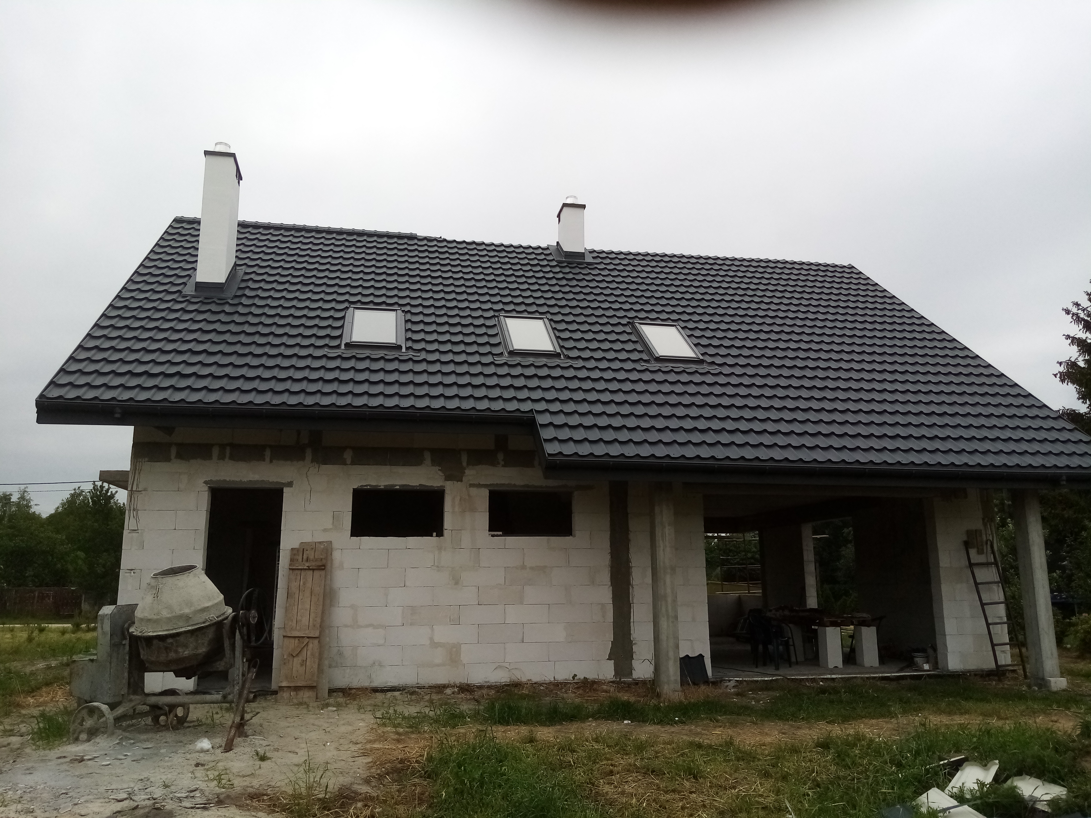 Surowy dom z ciemną dachówką, trzema oknami dachowymi i białymi kominami, betonowa mieszarka stoi przed budynkiem, widoczne niezabezpieczone otwory okienne.