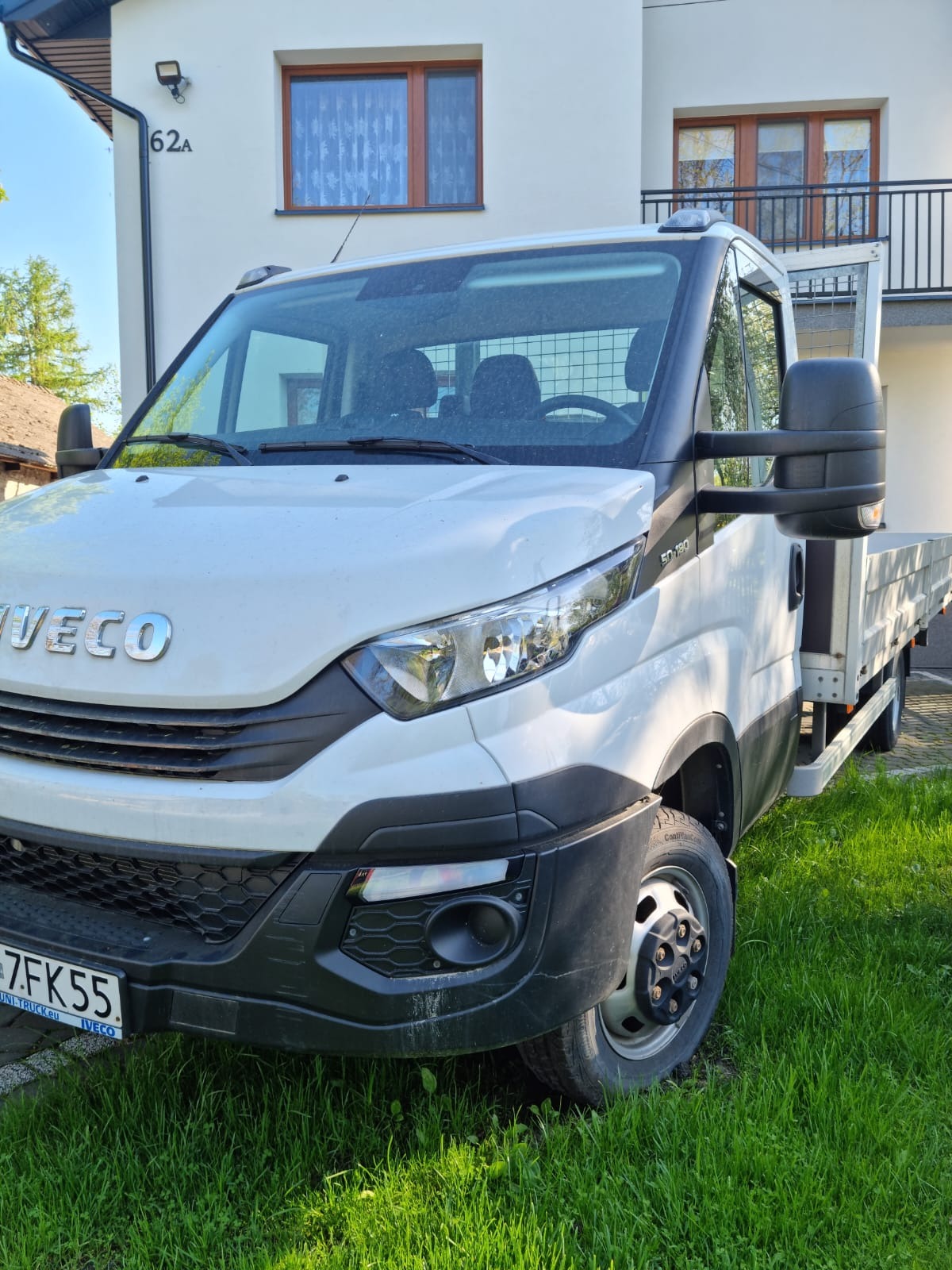 Biały samochód dostawczy Iveco z otwartą skrzynią ładunkową, zaparkowany na trawie przed budynkiem z numerem 62A.
