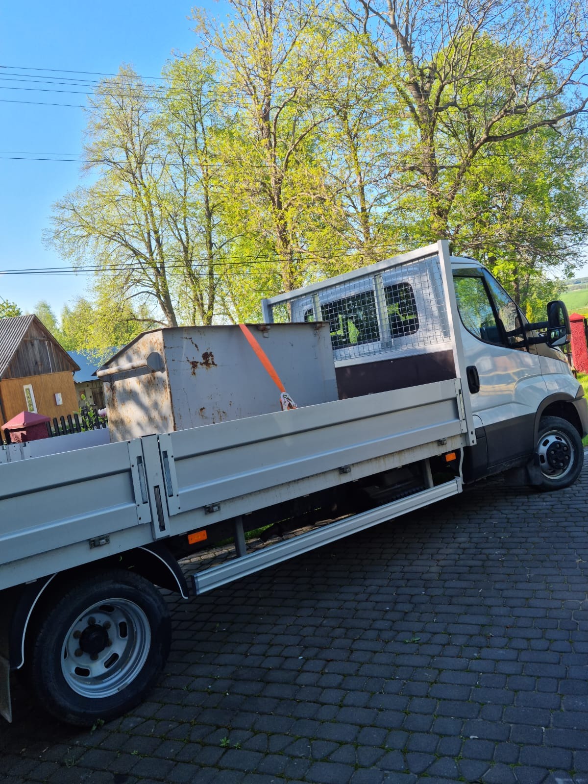 Biały bus z otwartą skrzynią ładunkową, zabezpieczony pomarańczowym pasem transportowym, przewożący rdzawy, metalowy kontener na tle zieleni drzew i błękitnego nieba.