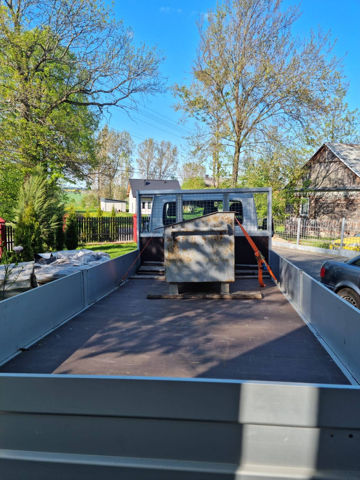 Pusta platforma transportowa z zabezpieczonym, rdzewiejącym metalowym elementem, otoczona zielenią i zabudowaniami.