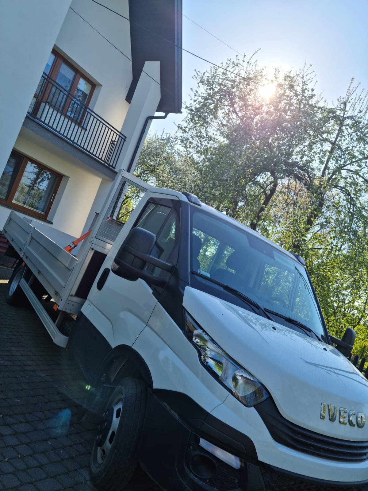 Biały samochód dostawczy Iveco z otwartą skrzynią ładunkową zaparkowany na brukowanej nawierzchni przed nowoczesnym domem z balkonem, w tle kwitnące drzewa i błękitne niebo z promieniami słońca.