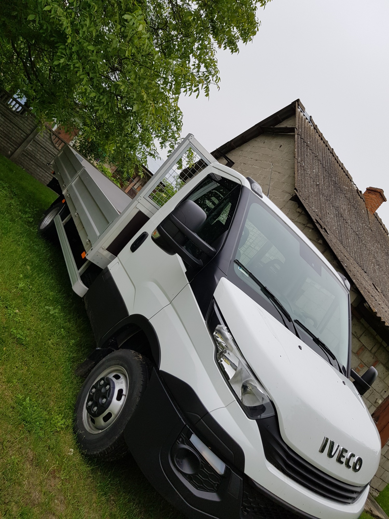 Biały samochód dostawczy Iveco z otwartą skrzynią ładunkową na tle zielonej trawy i starego domu z szarą dachówką.