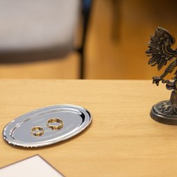 Niebiańska Fota - Dwa złote obrączki na srebrnej tacy z grawerem Urzędu Stanu Cywilnego, obok figurka orła na drewnianym stole, symbolizujące zawarcie związku małżeńskiego.