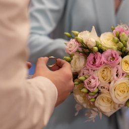 Niebiańska Fota - Dłoń trzyma złotą obrączkę ślubną na tle bukietu z różowych i kremowych kwiatów, z delikatnym logo fotografa w prawym górnym rogu.