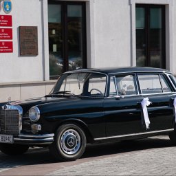 Niebiańska Fota - Czarny, klasyczny Mercedes z białymi wstążkami ślubnymi, zaparkowany przed budynkiem Urzędu Miasta w Serocku w słoneczny dzień. Eleganckie auto do ślubu.