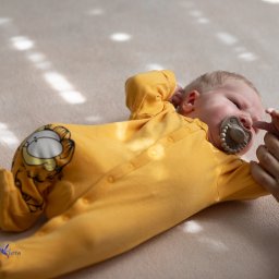 Niebiańska Fota - Leżący noworodek w żółtym pajacyku z Garfieldem, trzymający palec dorosłej osoby. Stonowana kolorystyka, naturalne światło, delikatne cienie.