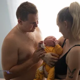 Rodzinne ujęcie: rodzice z noworodkiem w żółtym ubranku, tata trzyma smoczek, mama w czarnym biustonoszu, delikatne światło, intymna atmosfera.