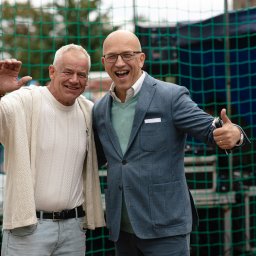 Niebiańska Fota - Uśmiechnięci mężczyźni pozują na tle siatki. Jeden w swetrze macha ręką, drugi w marynarce pokazuje kciuk w górę. Zdjęcie z eventu.