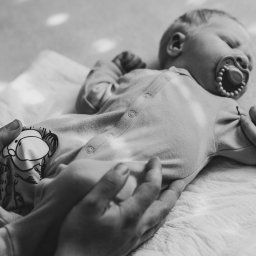 Niebiańska Fota - Czarno-białe zbliżenie śpiącego noworodka z smoczkiem, trzymanego w dłoniach rodziców na białym tle. Ubranko z motywem słonia.