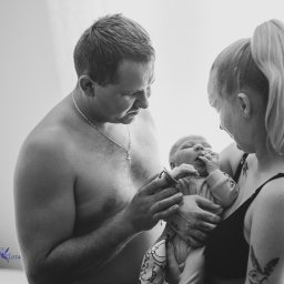 Niebiańska Fota - Czarno-białe zdjęcie rodziców z noworodkiem. Ojciec trzyma smoczek, matka przytula dziecko. Widoczne tatuaże na ciele matki. Minimalistyczne tło.
