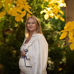 Niebiańska Fota - Portret kobiety w ciąży, w jesiennej scenerii. Żółte liście i zielone krzewy tworzą naturalne tło. Kobieta ubrana w jasny płaszcz, delikatny uśmiech.