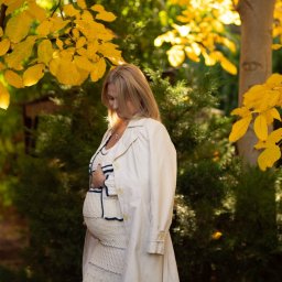 Niebiańska Fota - Kobieta w ciąży w kremowym płaszczu, pozuje na tle jesiennych drzew z żółtymi liśćmi. Delikatne światło podkreśla naturalność sesji ciążowej.