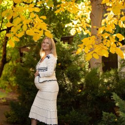 Niebiańska Fota - Uśmiechnięta, przyszła mama w dzierganej sukience pozuje na tle jesiennych drzew z żółtymi liśćmi. Romantyczna sesja ciążowa w plenerze, ciepłe światło.